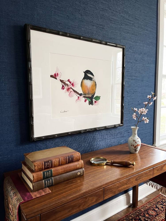 An original watercolor painting of a chickadee bird perched on a flowering branch, displayed on a textured blue wall above a wooden console table with books and a vase.
