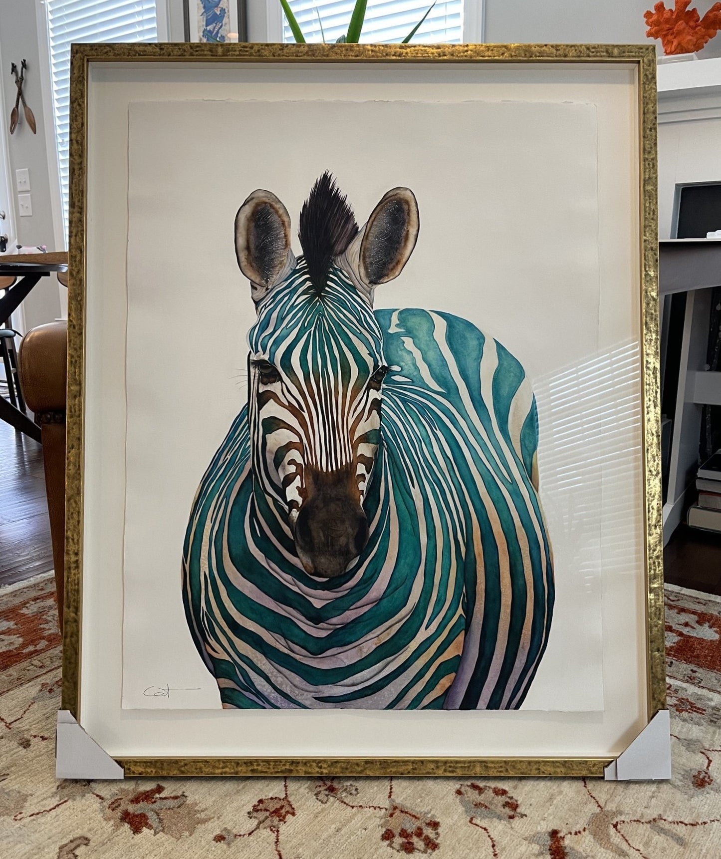A framed watercolor painting of Jade, featuring a stylized zebra with teal, green, and beige stripes on a white background, enclosed in a gold frame and leaning on a patterned rug in an indoor setting.
