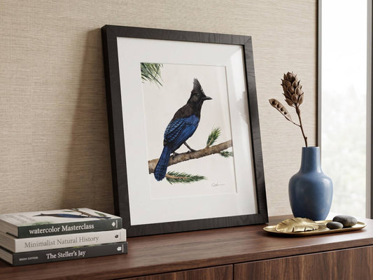 A framed watercolor art print of a Steller's jay perched on a branch, styled on a wooden console table next to a blue vase with dried stems and books.
