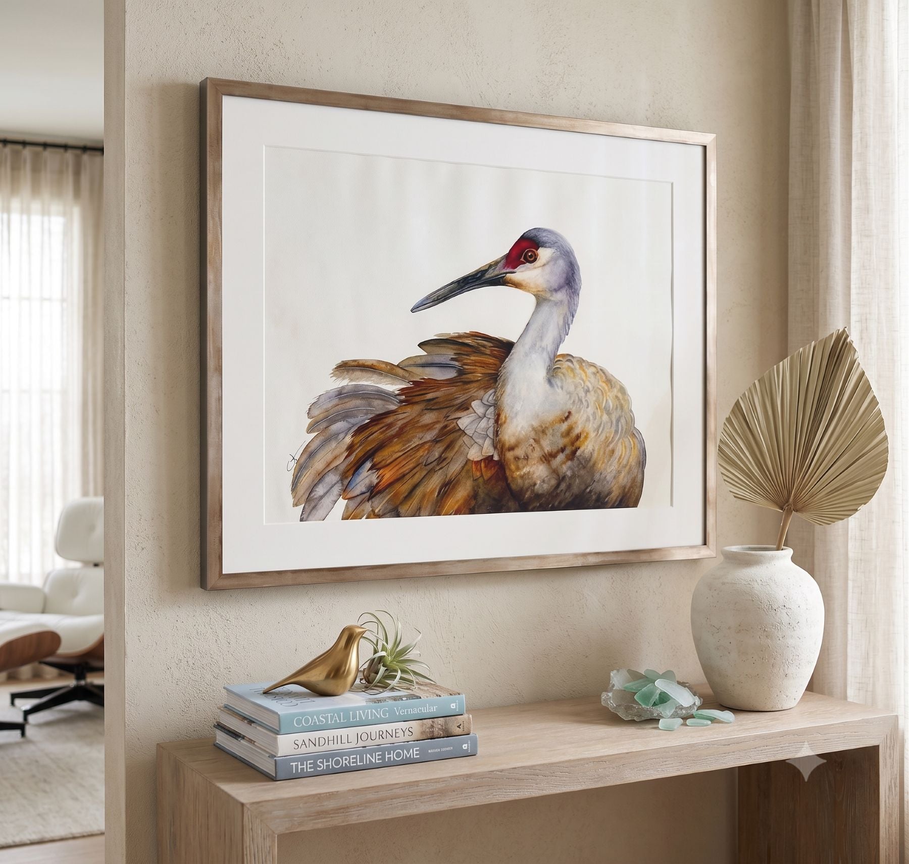 A framed watercolor painting of a sandhill crane by Vanessa Thorman hangs on a beige wall above a wooden console table in a styled interior. 
