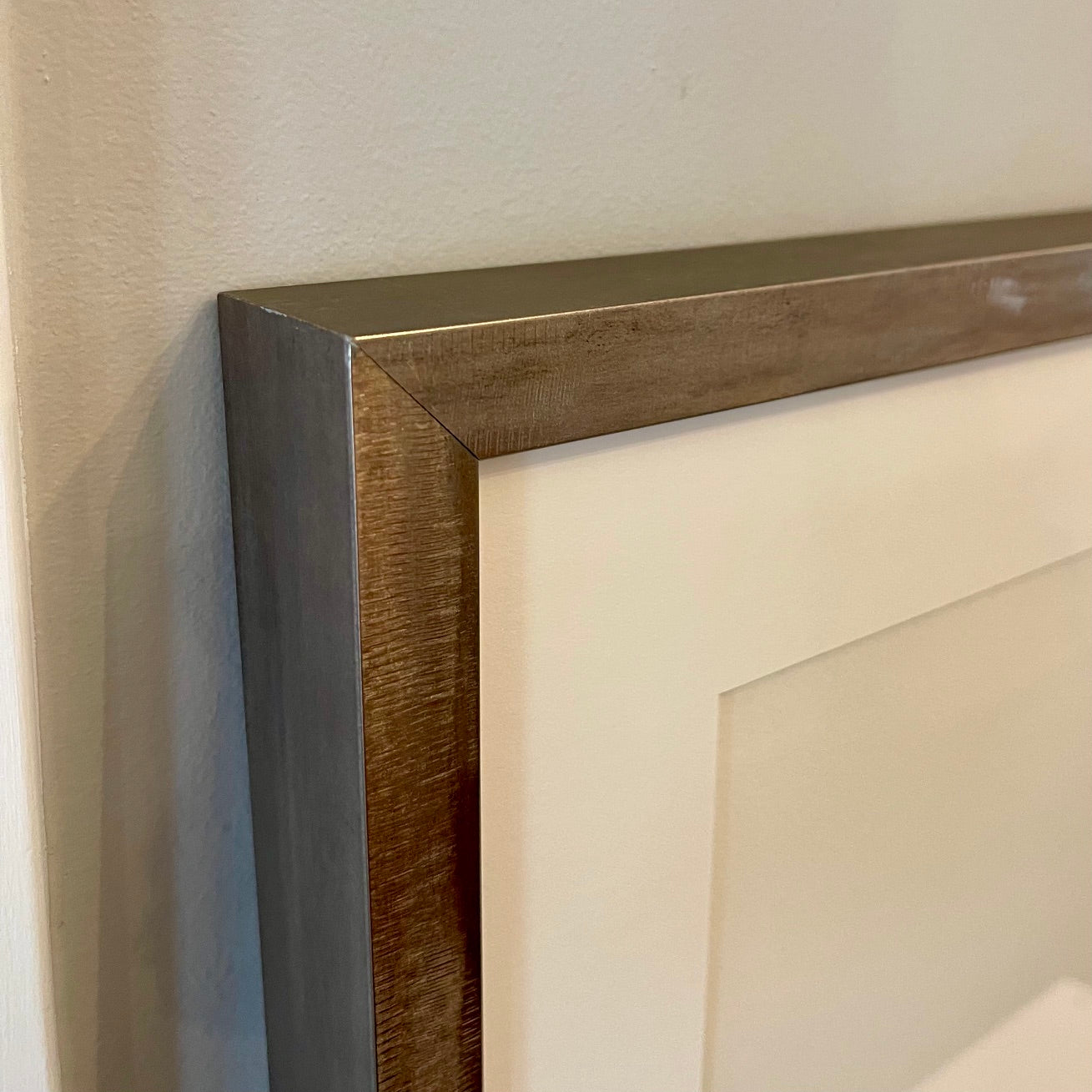Close-up of a framed wall art piece showing the corner joint, with a dark brown wood frame, a brushed silver metal inner frame, and an off-white mat against a beige wall.