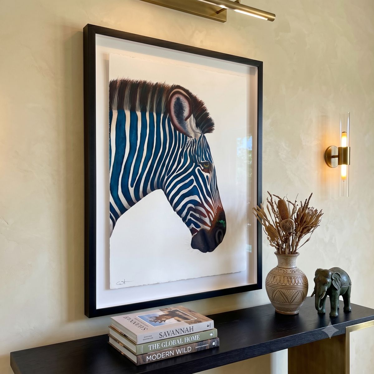 A framed watercolor of Ceasar, a blue zebra portrait, hangs on a beige wall above a dark console table. 