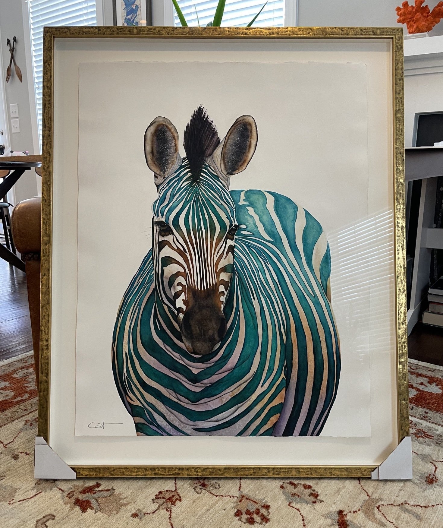 A framed watercolor painting of Jade, featuring a stylized zebra with teal, green, and beige stripes on a white background, enclosed in a gold frame and leaning on a patterned rug in an indoor setting.
