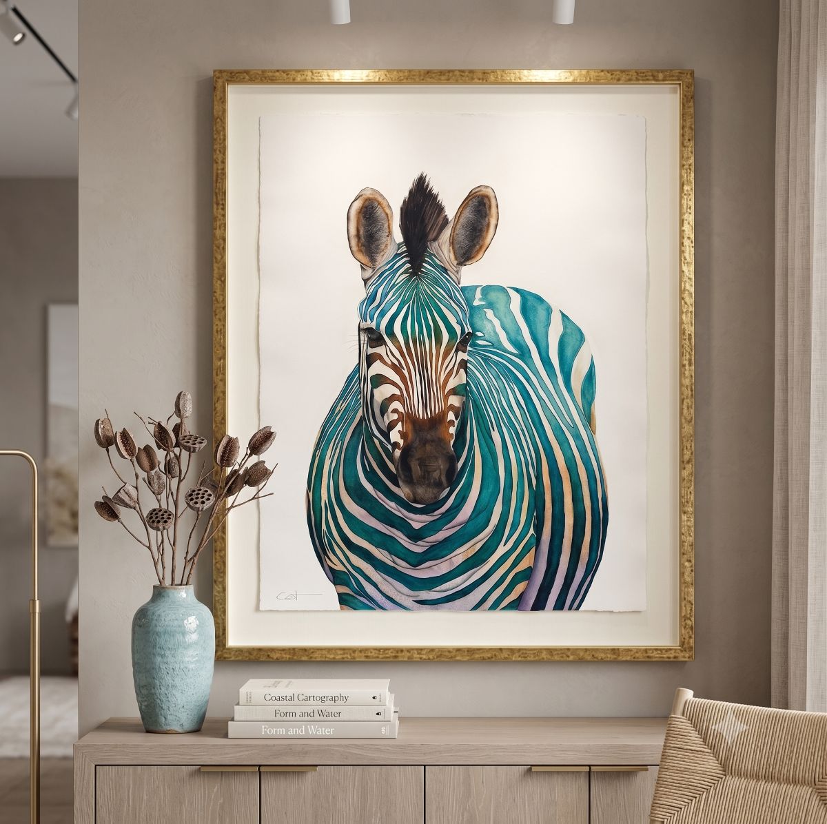 A framed watercolor painting of Jade, a zebra with teal, green, and white stripes, displayed in a gold frame on a beige wall above a light wood console table with a blue vase and books.